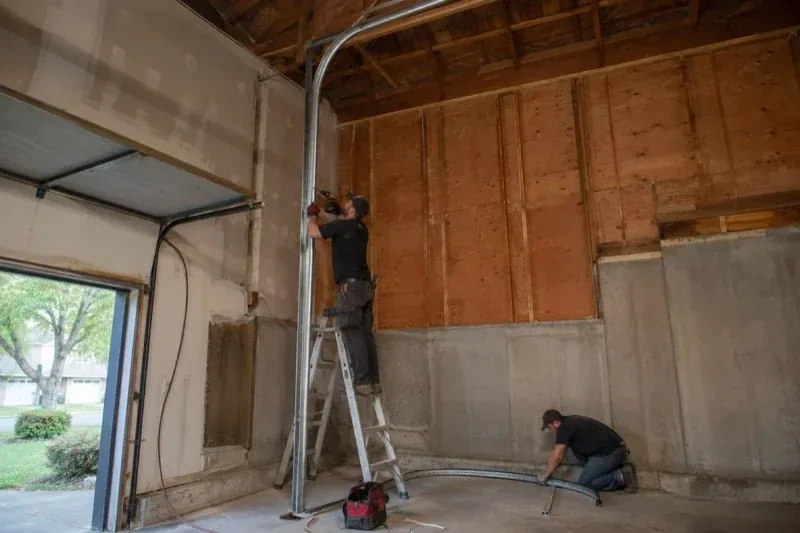 One person on a stepladder working beside a tall curved garage-door track while another kneels beside loose track sections in an unfinished garage. — High-Lift Garage Door Conversion, Mississauga